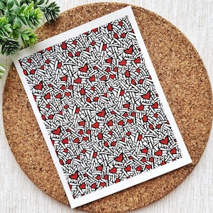 May include: A white card with a black and white pattern of lines and red hearts. The card is on a cork coaster.