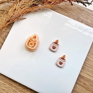 Pode incluir: Uma coleção de três pequenos objetos em forma de vela de cor clara com detalhes de coração. Um é um cortador de biscoitos e dois são velas em miniatura. Os objetos estão dispostos sobre uma superfície retangular branca com um fundo de madeira.
