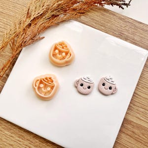 May include: A collection of craft supplies featuring a coffee cup theme. The image shows two coffee cup-shaped clay cutters and two small, pink, coffee cup-shaped clay charms with whipped cream and smiling faces. The items are on a white surface.