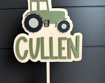 May include: A wooden cake topper featuring a layered design with a light green tractor at the top and the name CULLEN in dark green capital letters below. The entire piece is mounted on a thin wooden stick for insertion into a cake or dessert.