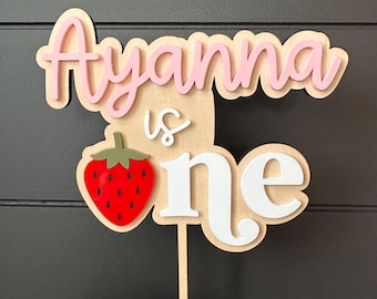 May include: A wooden cake topper featuring the text Ayanna is one in layered pink and white script and block lettering. A red strawberry with a green stem is attached to the left side. The entire design is mounted on a thin wooden stick.
