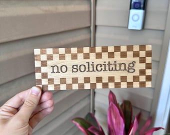 May include: Wooden sign with the words "no soliciting" in a cursive font. The sign has a checkerboard pattern border. The sign is held in front of a plant with pink and green leaves and a doorbell camera.