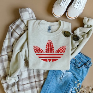 May include: A light beige sweatshirt with a red corn cob design resembling the Adidas logo. The sweatshirt is laid out on a plaid flannel shirt and blue jeans.