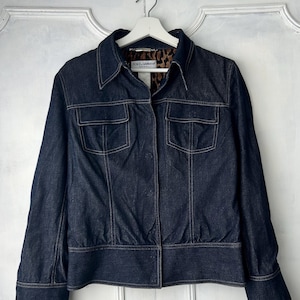 May include: A dark wash denim jacket with a classic collar, two chest pockets, and a fitted waist. The jacket features contrasting light stitching and a leopard print lining visible at the collar. The jacket is hanging on a white hanger.