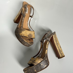 Pode incluir: Sandálias de plataforma com padrão de pele de cobra em tons de castanho, bege e bronzeado. As sandálias têm saltos grossos e tiras no tornozelo. A marca "PRADA" é visível na palmilha. Os saltos têm cerca de 10 cm.