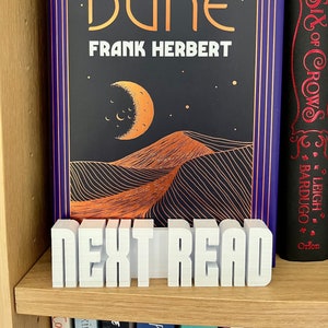 Könnte beinhalten: Ein Buch mit dem Titel "DUNE" von Frank Herbert, mit schwarzem Einband und goldfarbenem Schriftzug, das eine Wüstenlandschaft zeigt. Vor dem Buch steht ein weißer "NEXT READ" Buchständer auf einem Holzregal.