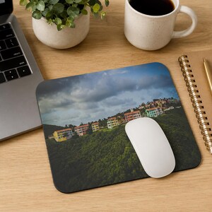 May include: A rectangular mousepad featuring a scenic landscape of colorful buildings on a green hillside under a cloudy sky. A white computer mouse rests on the mousepad, alongside a laptop, a coffee cup, and a small plant.