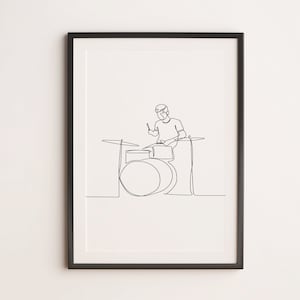 May include: A black and white line drawing of a person playing the drums. The drawing is framed in a black frame and is hanging on a white wall.