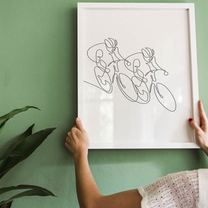 May include: A white framed print of a line drawing of two cyclists racing on bicycles. The print is hanging on a green wall.