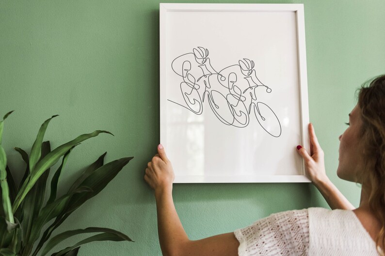 May include: A white framed print of a line drawing of two cyclists racing on bicycles. The print is hanging on a green wall.