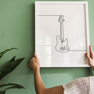 May include: A white framed print with a black line drawing of a bass guitar on a green wall. A person is hanging the print on the wall.