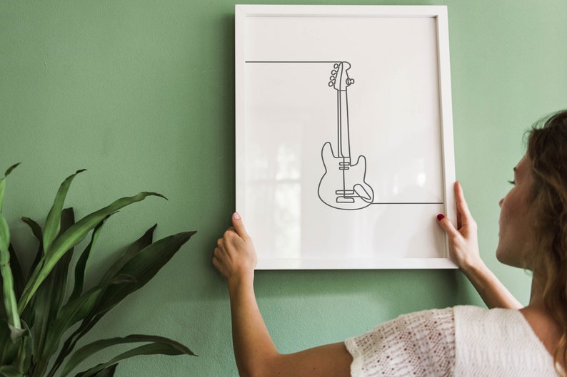 May include: A white framed print with a black line drawing of a bass guitar on a green wall. A person is hanging the print on the wall.