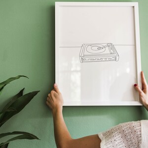 K&ouml;nnte beinhalten: Ein wei&szlig;er gerahmter Druck einer Strichzeichnung eines Plattenspielers an einer gr&uuml;nen Wand. Eine Person h&auml;ngt den Druck an die Wand.