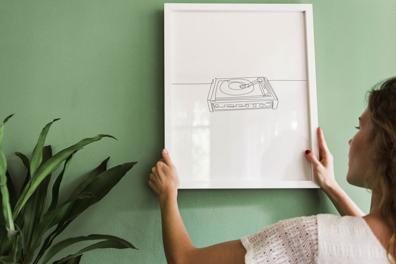 K&ouml;nnte beinhalten: Ein wei&szlig;er gerahmter Druck einer Strichzeichnung eines Plattenspielers an einer gr&uuml;nen Wand. Eine Person h&auml;ngt den Druck an die Wand.