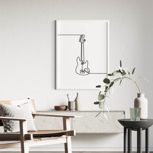 May include: A black and white line drawing of a bass guitar in a white frame hanging on a white wall. The frame is above a light brown wooden shelf with a vase and a bowl on it. A light brown chair with a white and black checkered pillow is in the foreground.
