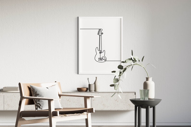 May include: A black and white line drawing of a bass guitar in a white frame hanging on a white wall. The frame is above a light brown wooden shelf with a vase and a bowl on it. A light brown chair with a white and black checkered pillow is in the foreground.