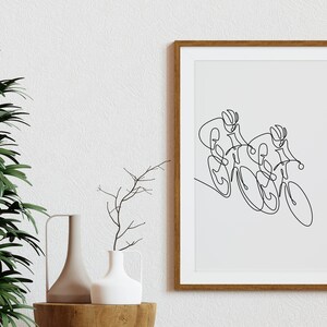 May include: A black and white line drawing of two cyclists racing on bicycles. The drawing is framed in a light brown wood frame and is hanging on a white wall.