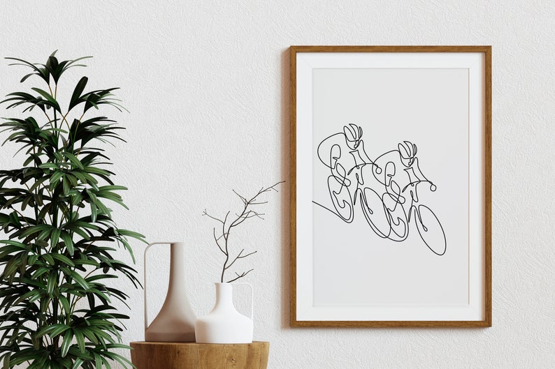 May include: A black and white line drawing of two cyclists racing on bicycles. The drawing is framed in a light brown wood frame and is hanging on a white wall.