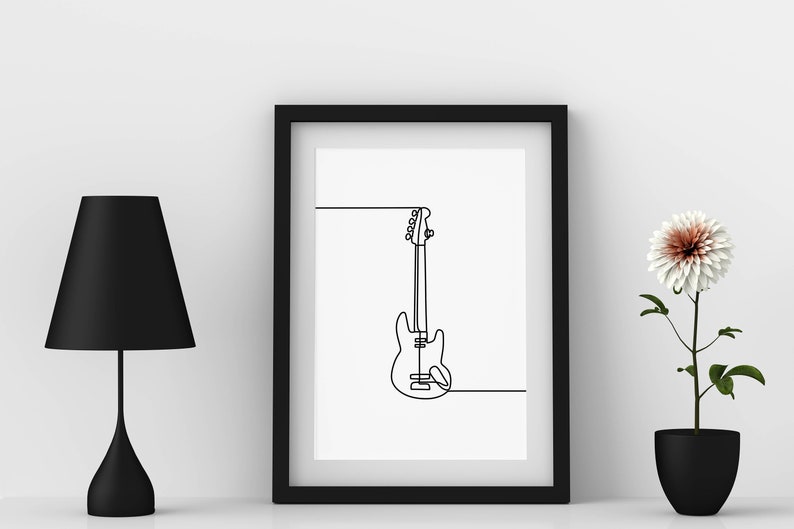 May include: A black framed print of a line drawing of a bass guitar hanging on a white wall. A black lamp and a potted white flower are on a white surface.