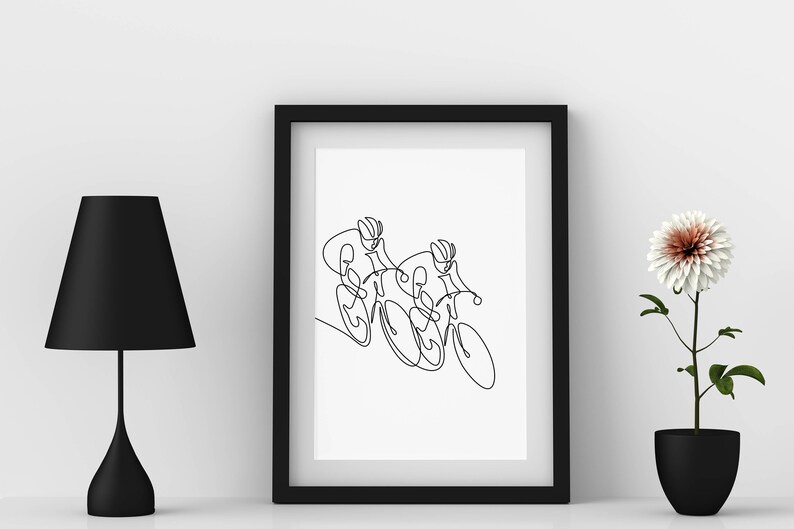 May include: A black framed print featuring a line drawing of two cyclists riding alongside each other. The print is displayed on a white wall, accompanied by a black lamp and a potted white flower.