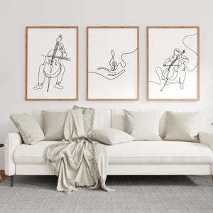 May include: Three framed art prints with line drawings of musical themes. The prints feature a cello player, a treble clef in a hand, and another cello player. The frames are a light brown color, and the art is displayed above a white sofa.