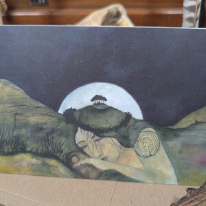 May include: A greeting card with an illustration of a person sleeping under a night sky. The artwork depicts a full moon, rolling hills, and a small tree. The colour palette includes muted greens, blues, and yellows, creating a tranquil scene.