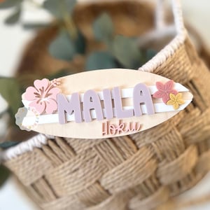 May include: A wooden surfboard-shaped sign with the name "Maila" in lavender letters and the word "Hoku" below. The sign is decorated with pink and yellow flower accents. The sign is set against a woven basket.