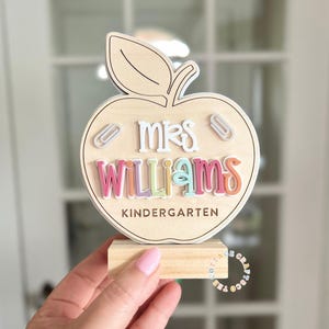 May include: Wooden apple-shaped teacher's desk decor with the name "Mrs. Williams" in colorful letters. The apple has a leaf and is mounted on a small wooden block. The word "Kindergarten" is also included.