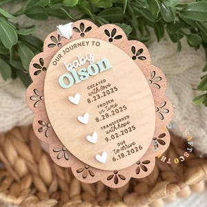 May include: An oval wooden ornament with a scalloped edge featuring the text OUR JOURNEY TO baby Olson, CREATED with love 8.23.2025, FROZEN in time 8.28.2025, TRANSFERRED with hope 9.02.2025, and DUE to arrive 6.18.2026 with four small white hearts.