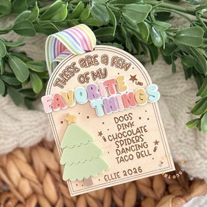 May include: An arched wooden ornament featuring the text "THESE ARE A FEW OF MY FAVORITE THINGS" in colorful 3D lettering. Below, a light green 3D Christmas tree sits next to a list: "DOGS, PINK, CHOCOLATE, SPIDERS, DANCING, TACO BELL" and "ELLIE 2026".