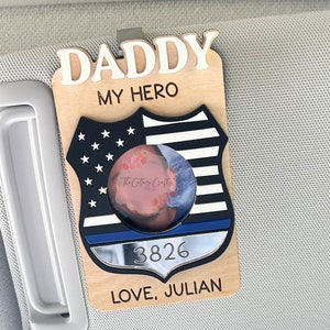 May include: Wooden car visor clip with a police badge design. The badge features a blue line, stars, and stripes, with the words "DADDY MY HERO" and "LOVE, JULIAN". Includes a photo and the number "3826".