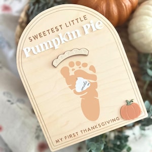May include: An arched wooden plaque featuring the text SWEETEST LITTLE Pumpkin Pie and MY FIRST THANKSGIVING. It displays a light orange footprint graphic with a white dollop shape labeled amelia 2024, a small arched accent, and a pumpkin icon.