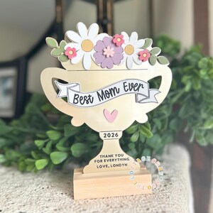 May include: A wooden trophy with a floral arrangement on top, featuring white daisies, pink and purple flowers, and green leaves. The trophy reads "Best Mom Ever" and "2026". The base says "THANK YOU FOR EVERYTHING LOVE, LONDYN".