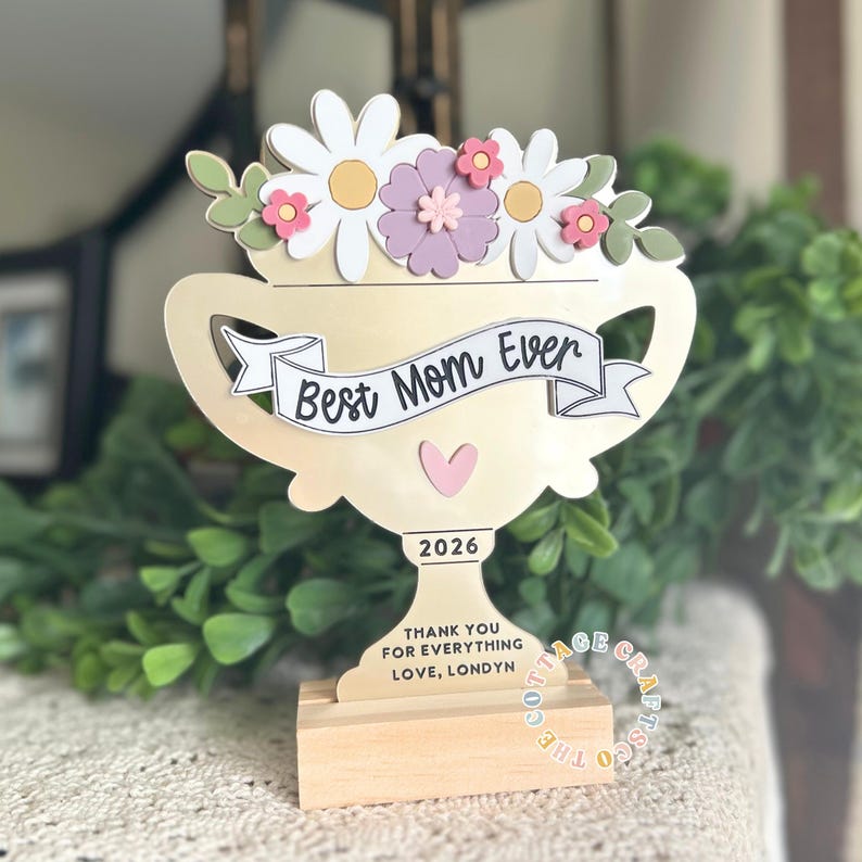 May include: A wooden trophy with a floral arrangement on top, featuring white daisies, pink and purple flowers, and green leaves. The trophy reads "Best Mom Ever" and "2026". The base says "THANK YOU FOR EVERYTHING LOVE, LONDYN".