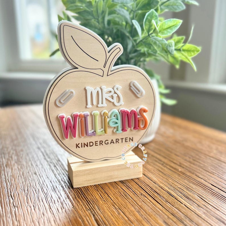May include: Wooden apple-shaped teacher desk sign with the name "Mrs. Williams" in colorful letters. The sign includes the word "Kindergarten" and two paperclip accents. The sign is displayed on a wooden stand.