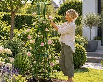 Op de afbeelding: Een hoge, donkergroene metalen obelisk-klimsteun met een uitlopende bovenkant, die een klimroos met lichtroze bloemen ondersteunt. De structuur bestaat uit verticale metalen staven en horizontale ringen voor stevige ondersteuning.
