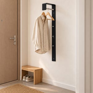 Pode incluir: Um cabideiro de parede vertical em metal preto com uma prateleira superior e uma barra horizontal para cabides. O suporte possui cinco pequenos ganchos prateados ao longo do comprimento. Uma camisa bege de manga comprida está pendurada.