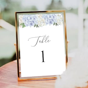 Pode incluir: Um cartão de número de mesa branco com moldura dourada. O cartão tem um design floral com hortênsias azuis e brancas. O texto no cartão diz "Mesa 1".