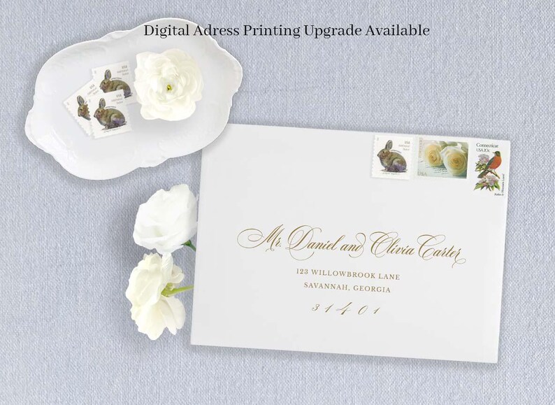 May include: A white envelope addressed to Mr. Daniel and Olivia Carter with gold calligraphy, featuring three postage stamps depicting a rabbit, roses, and a bird. A white scalloped dish holds additional rabbit stamps and white flowers.
