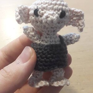 May include: A crocheted amigurumi doll of a house elf wearing a grey and white outfit. The doll has large ears and black eyes.