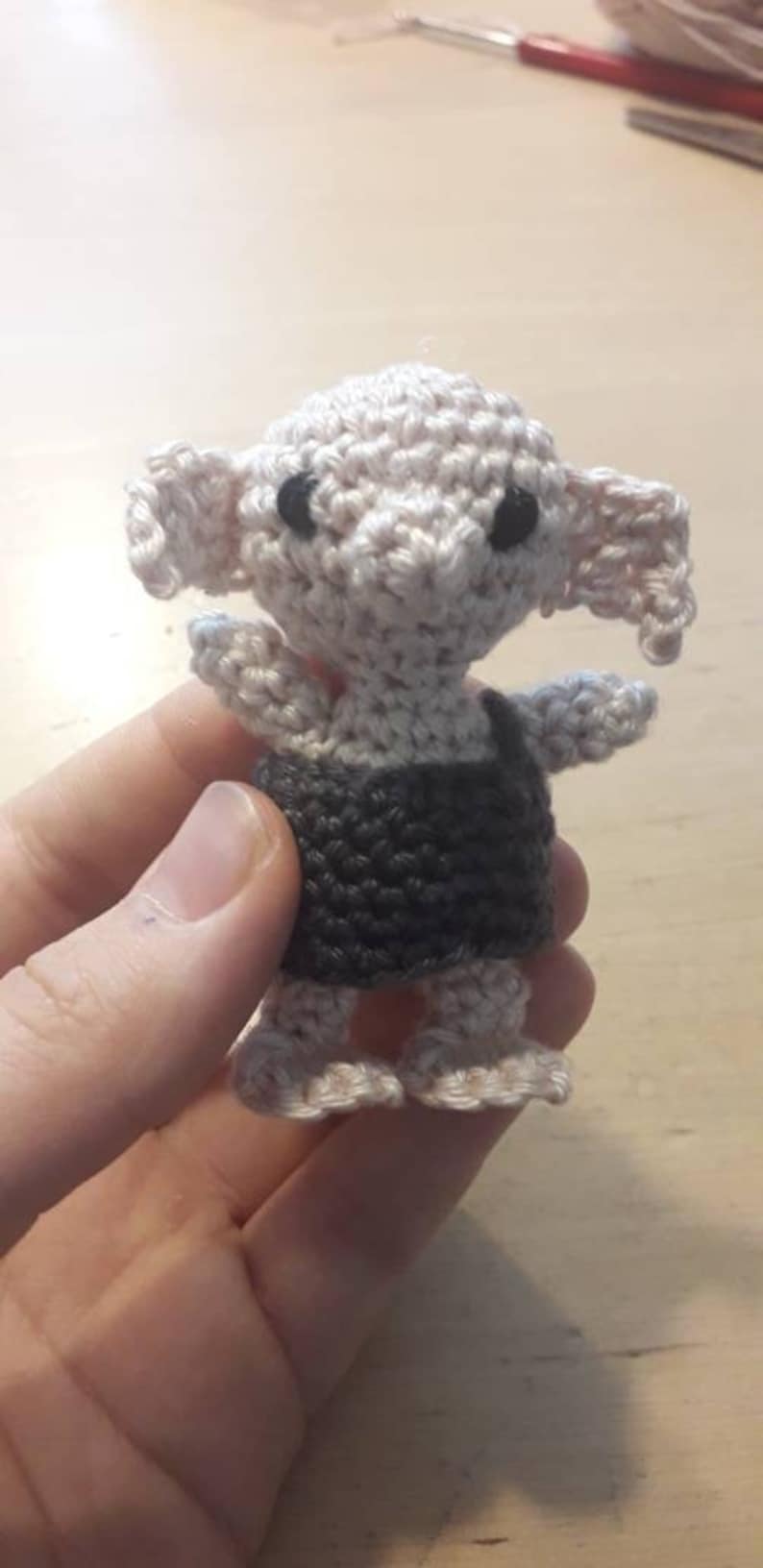 May include: A crocheted amigurumi doll of a house elf wearing a grey and white outfit. The doll has large ears and black eyes.