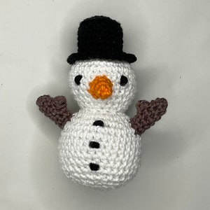 May include: A handmade crochet snowman with a black top hat, brown arms, and an orange carrot nose. The snowman has black button eyes and three black buttons down the front. The snowman is white.