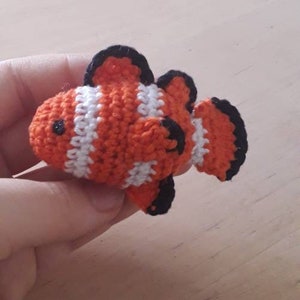 May include: A crocheted orange and white clownfish with black accents. The fish is small and has a detailed design.