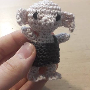 May include: A crocheted Dobby doll with a white body, black overalls, and black eyes. The doll is being held by a hand.
