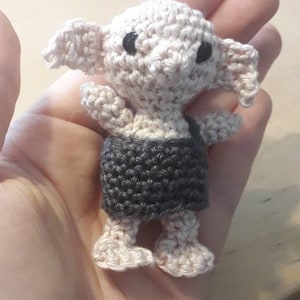 May include: A crocheted amigurumi doll of a house elf wearing a gray and white outfit. The doll has large ears and big eyes.