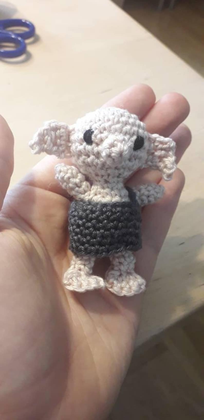 May include: A crocheted amigurumi doll of a house elf wearing a gray and white outfit. The doll has large ears and big eyes.