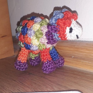 May include: A colorful crocheted sheep toy with a white face and purple legs. The sheep is made with a variety of colors, including red, orange, yellow, green, blue, and purple.