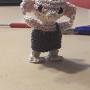May include: A crocheted Dobby doll with a gray and white outfit. The doll has large ears and black eyes.