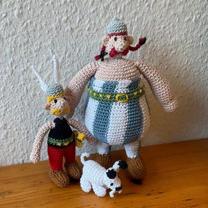 May include: Three handmade crochet dolls representing two human figures and a small dog. The smaller figure wears a winged helmet, black top, red pants, and brown boots. The larger figure has red braids, a blue and white striped outfit, and brown boots.