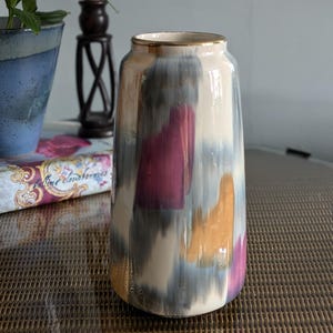 May include: A cylindrical ceramic vase featuring a cream-coloured base with abstract brushstrokes in shades of magenta, muted blue-grey, and golden yellow. The vase has a smooth, glossy finish and a thin, metallic gold rim around the top opening.